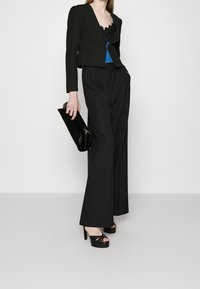 Černá zkrácená bunda přes modré tričko s krajkovým lemem, kombinovaná s černými kalhotami s vysokým pasem a širokými nohavicemi, obuté v černých sandálech na podpatku, a držící černou kabelku.