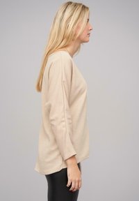 Beige long-sleeve top met een ronde hals, relaxte pasvorm en iets verlaagde schouders. Zachte stof met minimale textuur. Gecombineerd met zwarte leggings.
