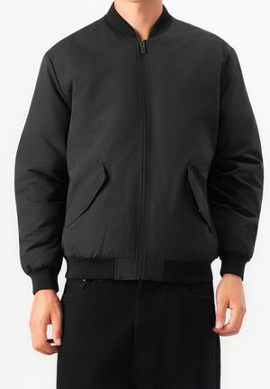 Schwarze gesteppte Bomberjacke mit Reißverschluss, geripptem Kragen, Bündchen und Saum, getragen über schwarzen Hosen, Hände entspannt an den Seiten.