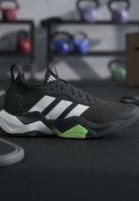 Chaussure de sport noire avec des bandes blanches et des accents verts sur le sol de la salle de sport, kettlebells et équipements d'entraînement flous en arrière-plan.
