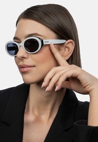 Aurinkolasit, joissa on soikeat mustat linssit, valkoinen kehys ja mustat yksityiskohdat. Malli koskettaa kehystä ja käyttää mustaa bleiseriä. Sileä pinta.