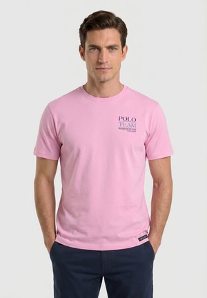 Hombre con camiseta de manga corta de color rosa claro con el texto "POLO TEAM" en el pecho y pantalones azul oscuro, de pie con las manos en los bolsillos.