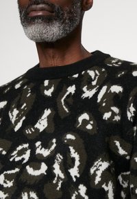 Pull en tricot avec une base noire et un imprimé léopard abstrait en vert olive et blanc. Encolure ronde classique et tissu texturé.