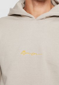 Detailní záběr na osobu v béžové mikině s kapucí, na které je žlutě vyšívané slovo "Člen" na hrudi.