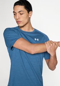 Μπλε αθλητική κοντομάνικη μπλούζα με υφασμάτινη υφή. Λευκό λογότυπο στην αριστερή πλευρά του στήθους. Το μοντέλο επεκτείνει το χέρι, παρουσιάζοντας τη εφαρμογή και το σχεδιασμό.