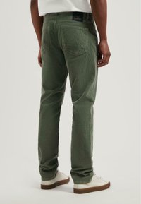 Pantalones verdes texturizados con corte recto, bolsillos traseros y una etiqueta de logo en la cintura, combinados con zapatillas blancas y suelas marrones.
