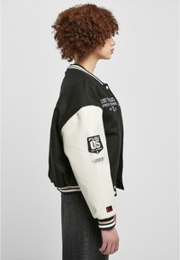 Čierno-biely varsity bunda s rebrovanými manžetami, obsahujúca nášivky FUBU a vyšívaný text. Jemný materiál, voľný strih a zapínanie na gombíky.