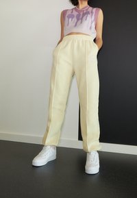 Krémové volné tepláky s bočními pruhy a vyšitým logem. Nejlépe ladí s tílkem z pleteniny v levandulové a růžové barvě.