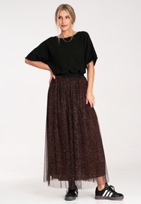 Czarna krótka bluzka zestawiona z długą, przezroczystą, brązową spódnicą z delikatnym wzorem. Modelka nosi czarne sneakersy z białymi paskami.