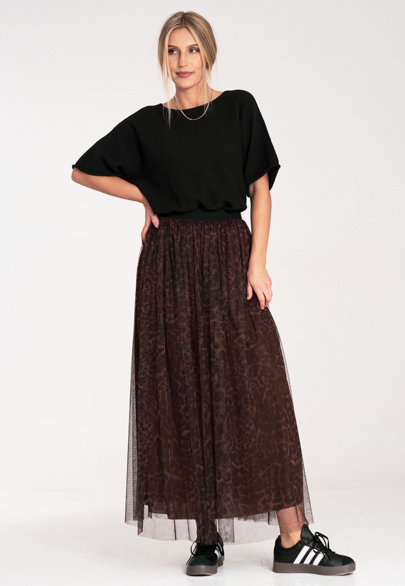 Czarna krótka bluzka zestawiona z długą, przezroczystą, brązową spódnicą z delikatnym wzorem. Modelka nosi czarne sneakersy z białymi paskami.