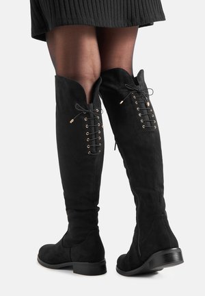 Botas negras hasta la rodilla con detalle de cordones en la parte trasera y cremallera lateral, combinadas con medias negras transparentes y una falda negra de canalé.