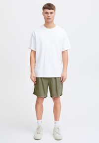 Tricou alb de bumbac, mâneci scurte, asortat cu pantaloni scurți texturați verde măsliniu și adidași bej, stând pe un fundal simplu.