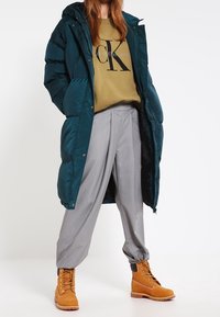 Giacca a vento teal con cappuccio, felpa khaki con logo, pantaloni grigi ampi e stivali beige con lacci e suola testurizzata.