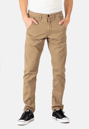 Pantalones de algodón beige con corte slim, cremallera con botón y bolsillos laterales, combinados con zapatillas negras con suelas de goma blancas.
