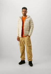 Beige dunjakke, orange skjorte, khaki cargobukser med lommer, sorte sko, model stående mod en ensfarvet baggrund.