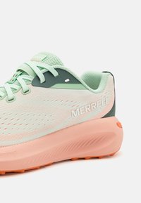 Zapatilla de running de malla verde claro con acentos en melocotón, que cuenta con una suela texturizada, talón reforzado y el nombre de la marca "MERRELL" en blanco.