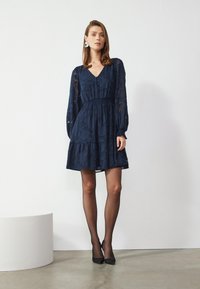 Robe bleu marine avec des manches longues transparentes, un col en V et une taille froncée. Motif floral texturé avec une jupe à volants. Escarpins noirs et collants.
