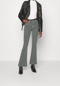 Zwarte leren bikerjack over een witte coltrui, gecombineerd met groene flared jeans en zwarte gestileerde laarzen met puntige neuzen.