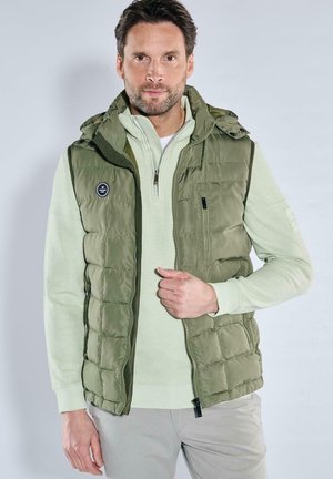 Green padded vest with a hood, featuring a zip closure and a small logo patch. Worn over a light green pullover. Light background.