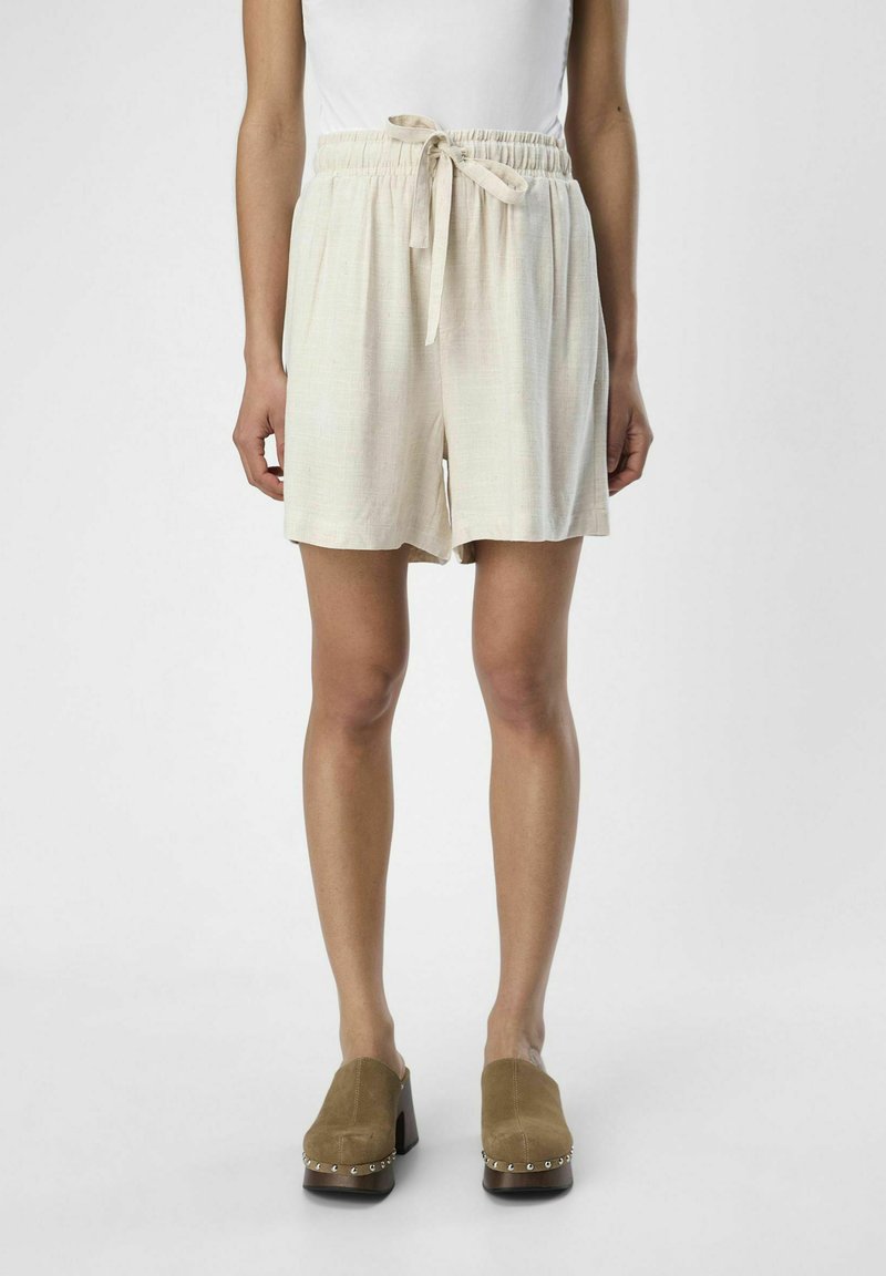 Lichtbeige, middellange shorts met een elastische tailleband en strikdetail. Glad, textuurstof. Gecombineerd met bruine heeled mules.