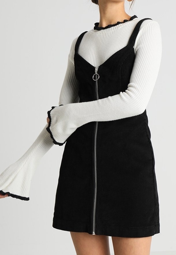 Svart kordflamsklänning med dragkedja fram, i kombination med en vit ribbad långärmad topp med trumpetärmar och scalloperade kanter.