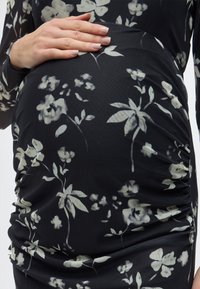 Μαύρο φόρεμα με λουλουδάτο μοτίβο σε λευκά λουλούδια, με ρυτιδωτό σχέδιο και απαλό, ελαστικό ύφασμα. Χέρι τοποθετημένο σε στρογγυλεμένη κοιλιά.