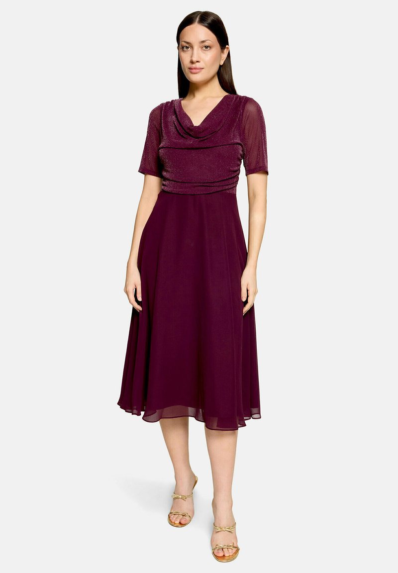 Femme portant une robe bordeaux à manches longueur genou avec un col drapé et des sandales dorées à bout ouvert, debout devant un fond clair.