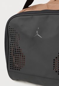 Mala de desporto cinza com um design perfurado, apresentando um topo textil bronze, fecho de correr preto e um logo Jumpman prateado na frente.