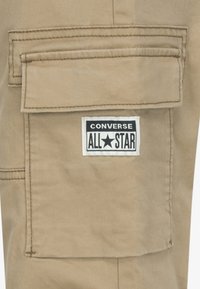 Bež hlače s stranskimi žepi in �črno-bela značko Converse All Star, ki je postavljena na vidnem mestu.