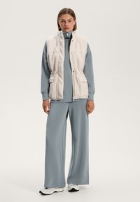 Umelý kožušinový vest v krémovej farbe, ktorý je oblečený cez svetlomodrú mikinu a široké nohavice, s praktickými vreckami a ležérnym športovým dizajnom.