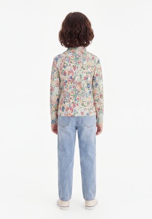 Enfant aux cheveux bruns bouclés portant une chemise fleurie à manches longues et un jean bleu clair, debout de dos sur un fond blanc.