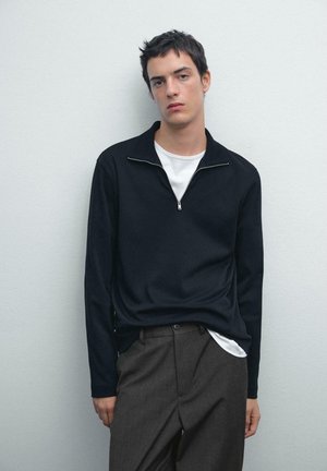 Black zip-up long-sleeve top over a white t-shirt, paired with gray trousers. Smooth texture, relaxed fit, minimalistic design.
