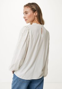 Blusa bianca con maniche a sbuffo, dettagli in pizzo e una piccola apertura a forma di chiave sul retro. Tessuto morbido con una texture liscia.