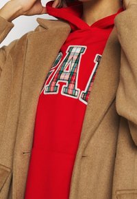 Abrigo de lana color camel sobre una sudadera con capucha roja con letras a cuadros, mezcla de algodón. El abrigo tiene un corte holgado con solapas y bolsillos laterales.