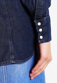 Mörkblå denimskjortärm med tre vita knappar på manschetten, med kontrasterande sömmar och rundad nederkant, hålls av en hand mot en ljus bakgrund.