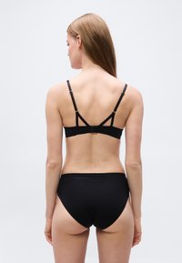 Γυναίκα με μακριά ανοιχτό καφέ μαλλιά, που φαίνεται από πίσω, φορώντας μαύρο σουτιέν με ρυθμιζόμενους ιμάντες και μαύρο ταίριαστο εσώρουχο σε απλό φόντο.