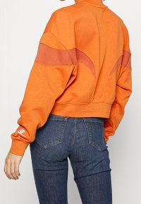 Orange kortärmad sweatshirt med kontrasterande texturerad paneldesign på axlarna, kombinerad med mörka jeans. Har ribbade muddar.