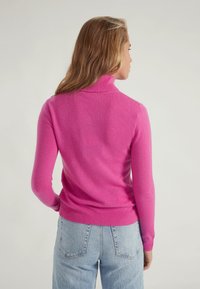 Maglione a collo alto fucsia in maglia con maniche lunghe e polsini a coste. Tessuto morbido, design aderente, abbinato a jeans azzurri a vita alta.