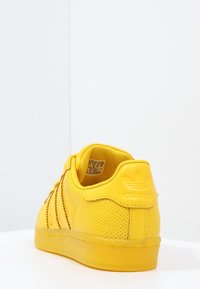 Zapatilla amarilla con superficie texturizada, perforaciones y tres rayas. Punta redondeada y suela de goma con logotipo en relieve en el talón.