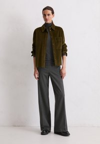 Chemise en velours côtelé couleur olive avec un bouton et une poche, superposée à un col roulé gris foncé. Pantalons à jambes larges gris et chaussures noires complètent le look.