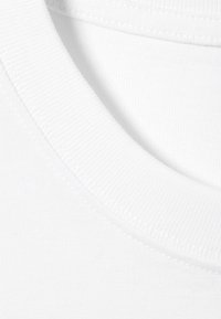 Hvit bomullsduk med ribbet rund hals. Glatt tekstur med søm detaljer langs kantene. Nærbilde av tekstilet.