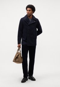 Marineblauer Doppeltspannmantel mit großen schwarzen Knöpfen, kombiniert mit dunklen Cordhosen und schwarzen Schuhen. Hält eine braune Ledertasche.