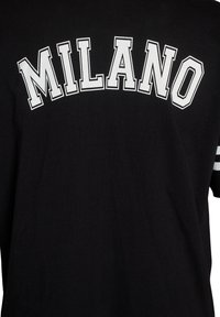 T-shirt noir avec "MILANO" en lettres capitales blanches et contournées, en gras, sur la poitrine, et deux bandes blanches sur la manche.