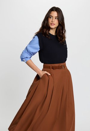 Woman with wavy brown hair wearing a black sleeveless sweater over a blue shirt and a belted brown maxi skirt, standing with hand in pocket.