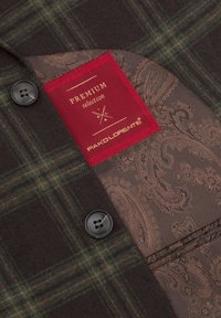 Brązowa i zielona tkanina w kratę z podszewką w wzór paisley. Posiada czerwoną metkę z napisem "PREMIUM selection" oraz czarny guzik.