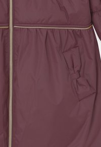 Veste rembourrée bordeaux avec une fermeture éclair dorée, taille élastiquée, et une poche avec un accent de nœud sur le côté gauche. Texture lisse et brillante.