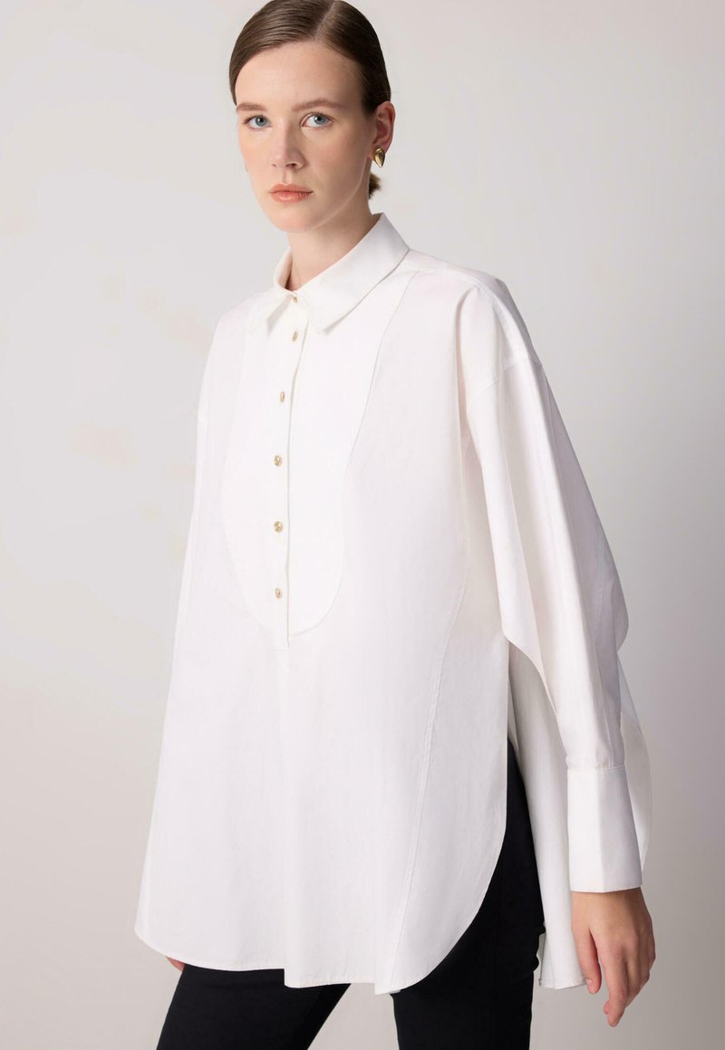 Chemise blanche surdimensionnée fabriquée dans un tissu doux. Dispose d'une fermeture à boutons à l'avant, d'un col évasé et de fentes latérales discrètes pour un mouvement accru.