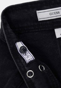 Fascia in denim nero con bottone nero, occhielli in metallo e etichette bianche con il nome del marchio "GUESS" cucite all'interno.