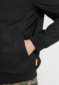 Svart jacka med en zip-ficka och orange accentetikett, med en strukturerad tyg och ribbade ärmslut för en åtsittande passform.