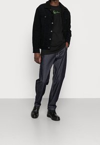 Giacca di velluto a coste nera con colletto in pelliccia sintetica, T-shirt nera con stampa verde, pantaloni di jeans scuri e stivali neri con lacci.